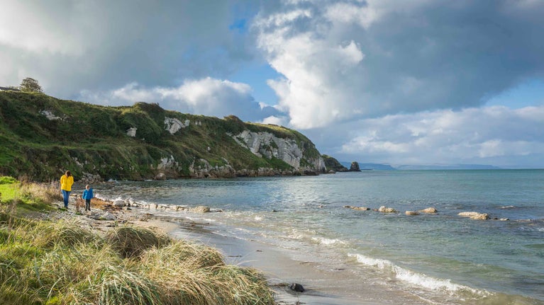 An adult and a child are walking along the grassy Portmuck coastline on the Islandmagee peninsula, County Antrim, with the clear blue-green water stretching out towards cliffs.